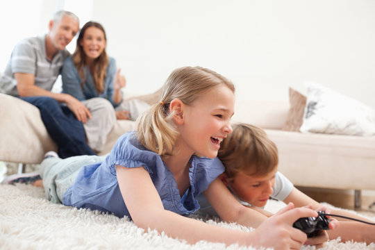 Siblings playing video games with their parents on the backgroun