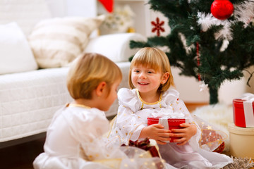 Two twins girl sitting with presents near Christmas tree