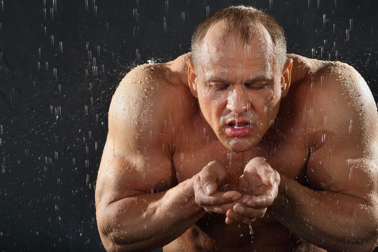 Undressed tanned bodybuilder in rain drinks water from hands