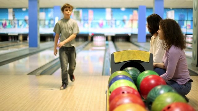 Parents sit and watch boy throws bowling ball to beat skittles