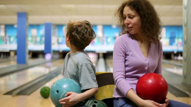 Mom and son hold bowling balls in hands and talk in bright club