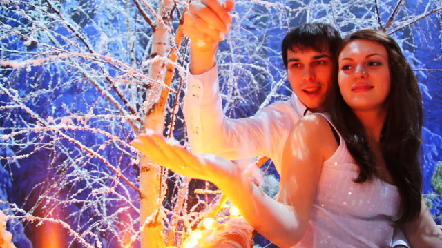 couple sits on lighted balcony in snowy birch forest at night