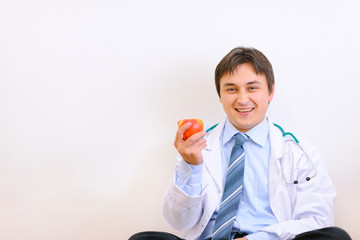 Smiling medical doctor sitting on floor and holding apple in han