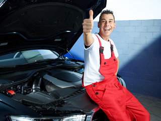 Happy apprentice in a garage sitting on a car shows thumb up