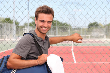 Man with sports bag stood by clay tennis court