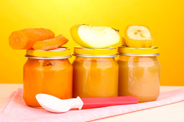 Jars of baby puree with spoon on napkin on yellow background