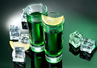 Two glasses of absinthe, lemon and ice on green background