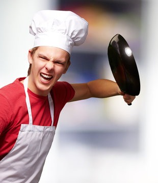 portrait of angry young cook man hitting with pan indoor