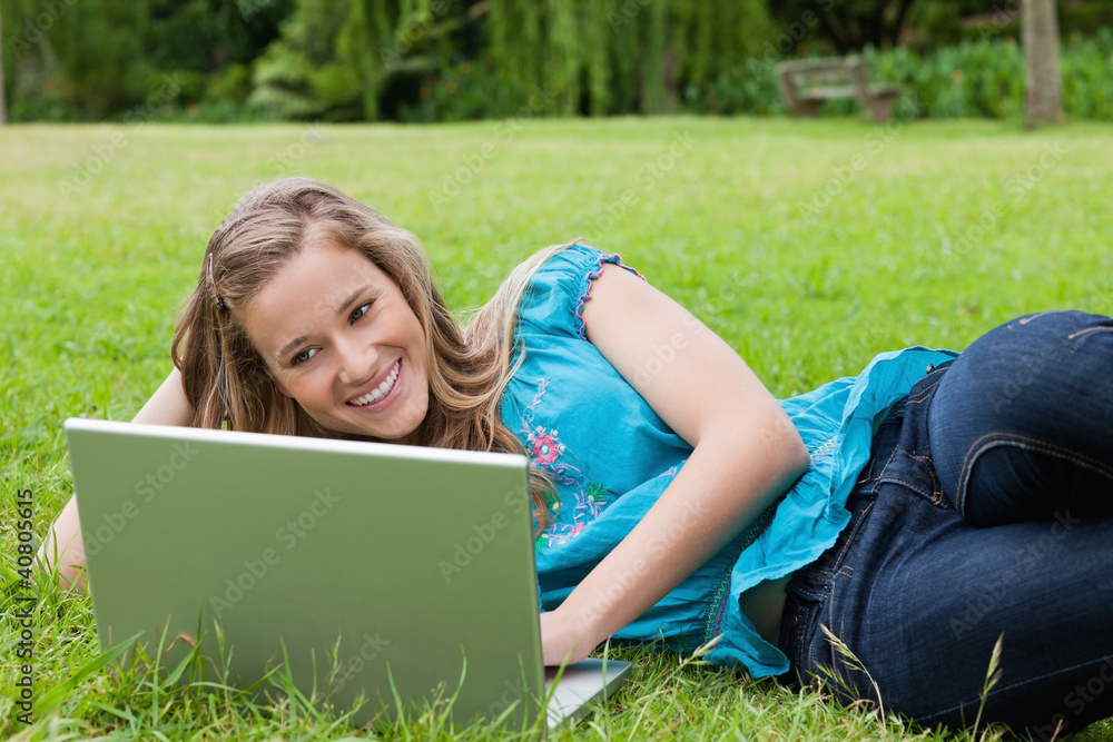 Poster young smiling girl lying on the side in a park while typing on h