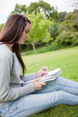 Obraz premium Side view of a young girl sitting on the grass while writing on