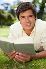 Man looking ahead while reading a book as he lies on grass