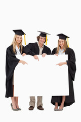 Three students in graduate robe holding and pointing a blank sig