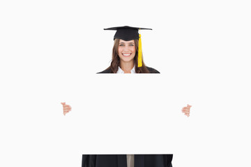 A woman in her graduation gown holds a blank sheet in front of h