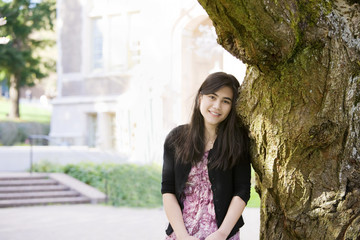 Teen girl leaning against trunk of large tree, smiling