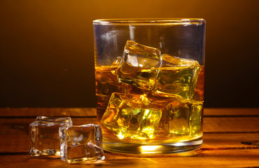 glass of whiskey and ice on wooden table on brown background