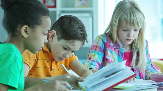 Three diverse girls and boy sitting at desk working on school task, checking information in books and writing notes. Perfect for back to school content