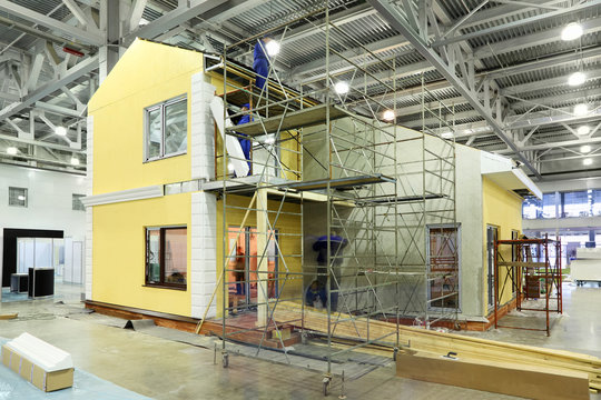 Team of builders build two-storey yellow cottage in large hall