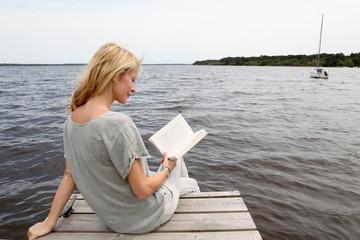 Woman reading book sitting on lake boardwalk