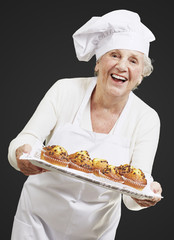 senior woman cook holding a tray with muffins against a black ba