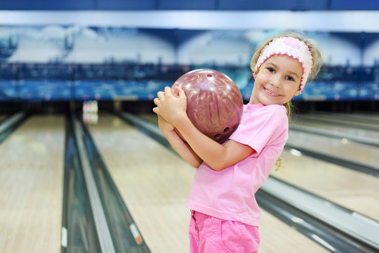 Little girl dressed in pink T-shirt holds ball in bowling club