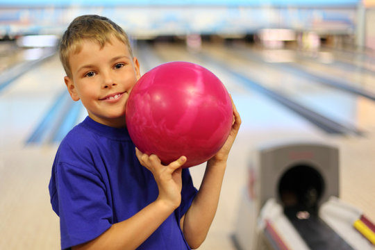 Smiling boy dressed in T-shirt holds pink ball in bowling club
