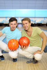 Two smiling men sit on floor with orange balls in bowling club;