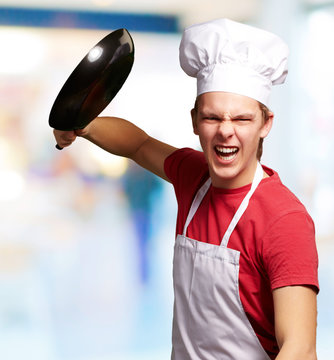 portrait of angry young cook man hitting with pan indoor