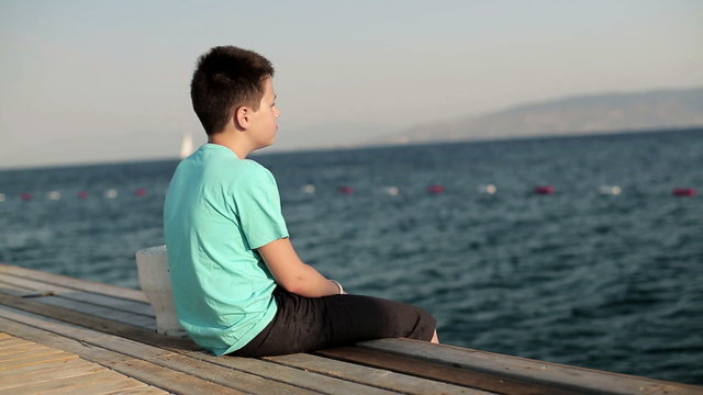 Lonely pensive boy sitting on wooden pier by the sea