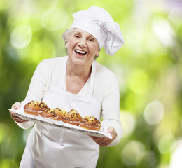 senior woman cook holding a tray with muffins against a nature b