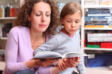 Daughter with her mother read book in room with bookshelves
