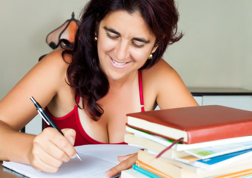 Hispanic Woman Writing With A Stack Of Books On Her Desk