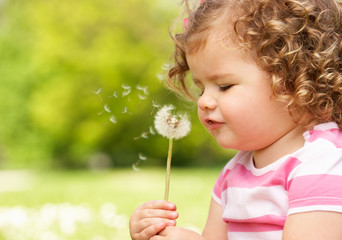Young Girl In Summer Dress Sitting In Field Blowing Dandelion