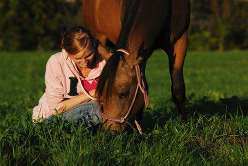 a girl sitting in the pasture beside her grazing horse - 44081003
