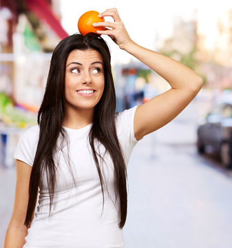 portrait of young woman holding orange on her head at city