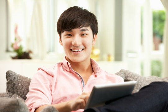 Young Chinese Man Using Digital Tablet Whilst Relaxing On Sofa