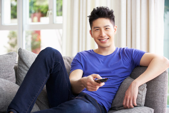 Young Chinese Man Sitting And Watching TV On Sofa At Home
