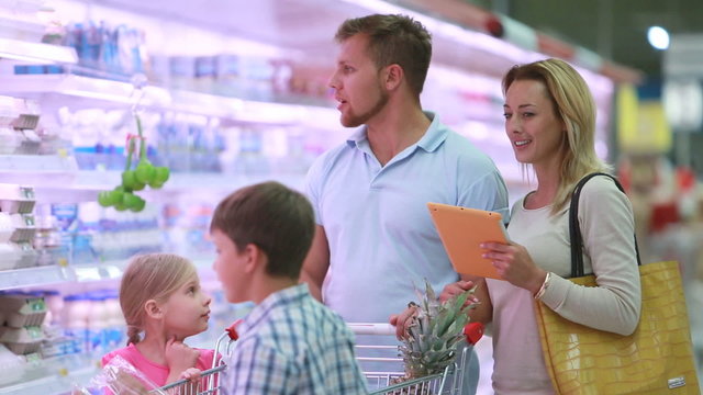 Family with two children choosing groceries in supermarket, checking shopping list on tablet. Suitable for retail, family life, budgeting and meal planning content