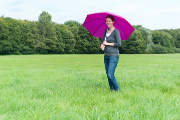 Frau mit braunen Haaren steht mit einem lila Sonnenschirm auf einer gr&uuml;nen Wiese
