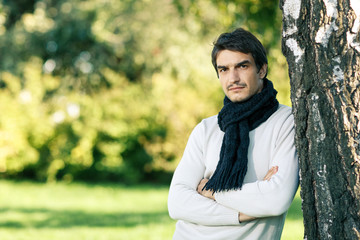 Man outdoors in fall clothing and scarf leaning on tree stem