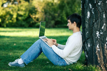 Young man sitting near the tree stem and using laptop