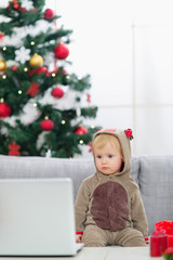 Baby in Christmas deer costume near Christmas tree looking in la