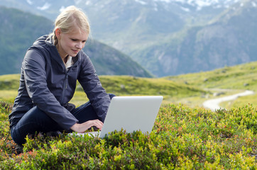 junge blone Frau sitzt mit Laptop in Bergwiese