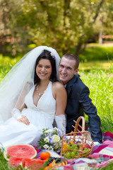 groom and the bride sit on a grass with a big basket with fruit