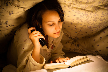 Serious Woman Reading a Book with Flashlight on Bed