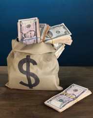 Bag with stacks of dollars on wooden table on blue background