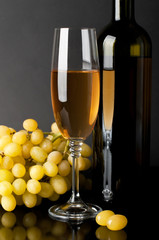 bottle and glass of white wine with grapes on dark background