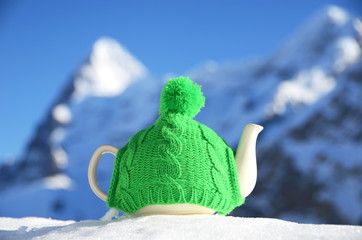 Tea pot in the knitted cap on the snow against mountain peak