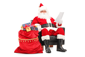 Santa Claus in armchair reading a letter next to a bag full of g