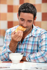 Smiling man eating croissant for breakfast with a magazine