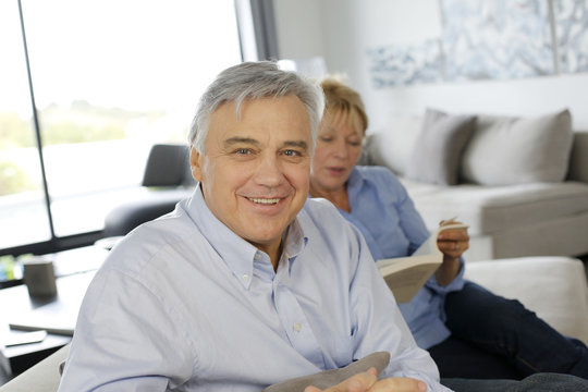 Smiling senior man sitting in couch, wife in background